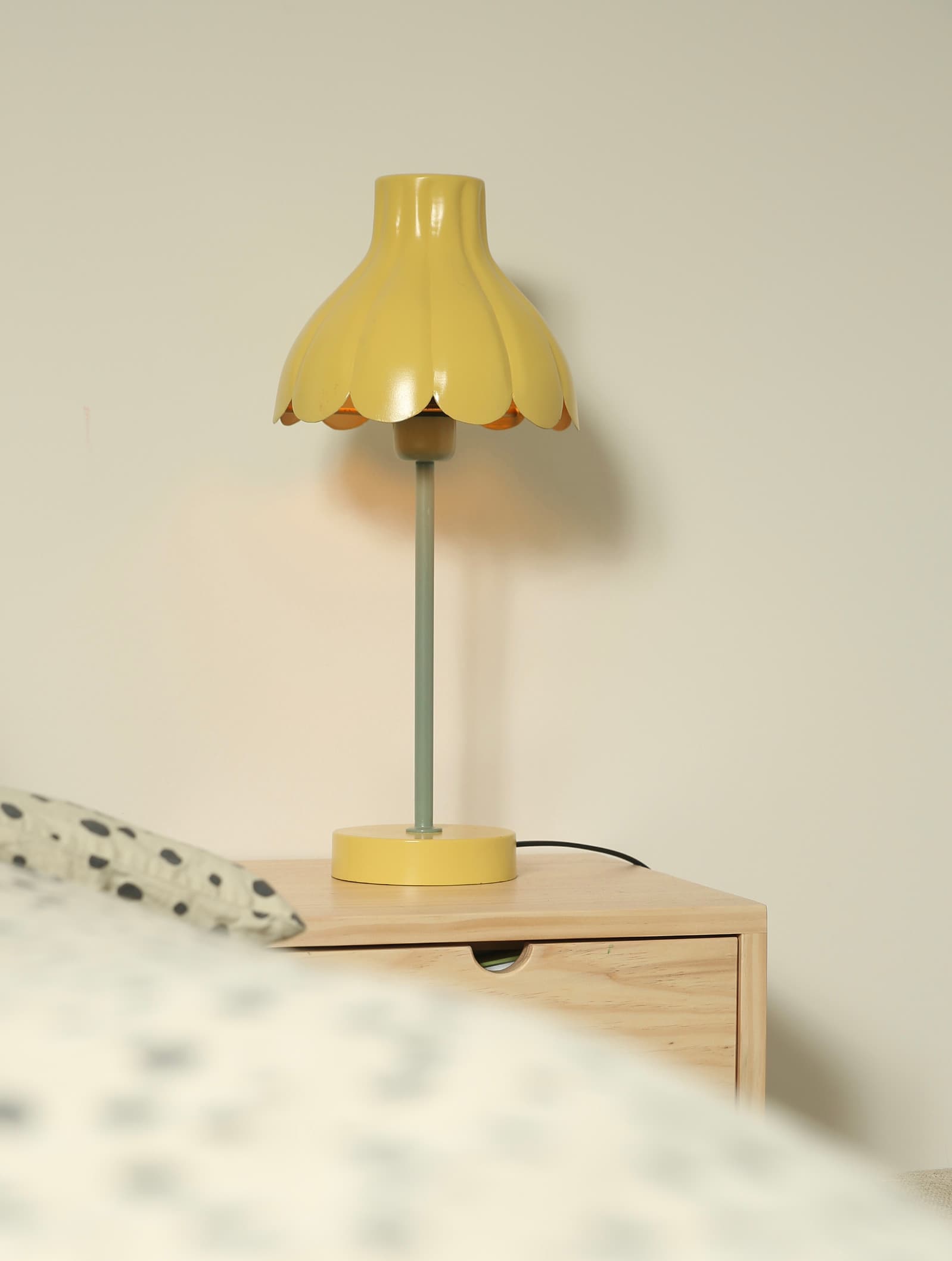 Gul lampa i metall med grön dekorativ stång på ett sidobord bredvid en säng.